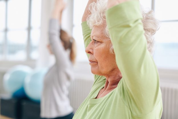 Older woman stretching up shoulder with younger woman next to her in class